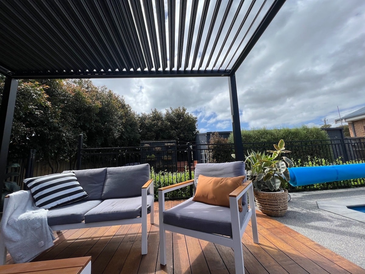 Poolside pergola with black louvre roof and outdoor lounge seating on timber deck, offering stylish shade and relaxation.