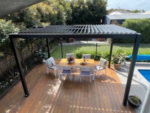 Elegant patio with louvered roof for pergola, dining table, chairs, and lush greenery.