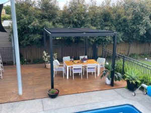 Minimalist front patio pergola with a sleek black frame, shading an outdoor dining area on a wooden deck.