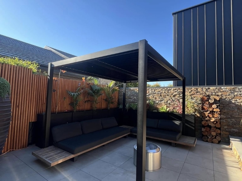 Best Pergola Material shown in modern black louvre pergola with outdoor lounge seating in compact courtyard.