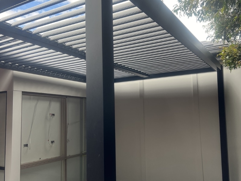 Corner Pergola Designs featuring black louvre pergola attached to house with enclosed white walls.