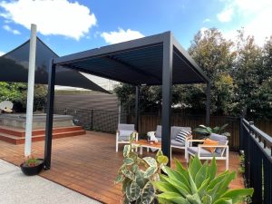 Modern pergola decking ideas with a black aluminium pergola, wooden deck, outdoor seating, and lush greenery for a cosy backyard retreat.