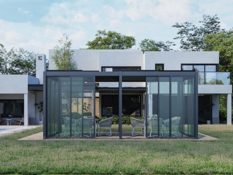 Pergola All Seasons shown as glass-enclosed outdoor structure providing year-round dining space beside modern home.