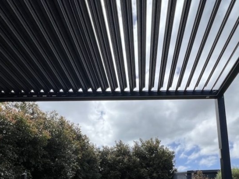 Pergola in rooftop with black louvered panels partially open against cloudy sky and surrounding trees.