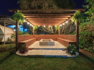 Cosy wooden pergola with built-in benches, pergola side panels, stone flooring and lush greenery in a backyard setting.