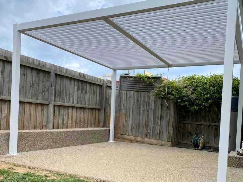 Adjustable pergola with white louvered roof installed over a concrete outdoor area beside timber fencing.