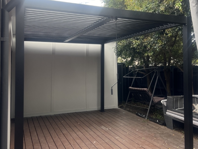 Corner pergola with sides featuring black frame and louvered roof installed over timber deck.