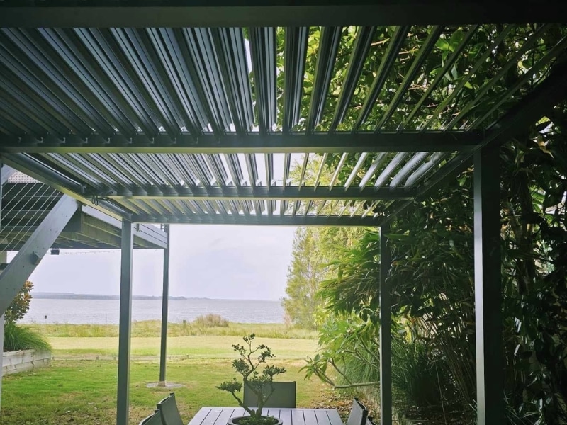 Parts of a pergola shown in a metal louvre structure overlooking a lakeside garden.