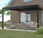 Modern wall-mounted manual louvered pergola with sleek black frame over a cozy outdoor lounge area featuring light wooden furniture and a brick house backdrop.