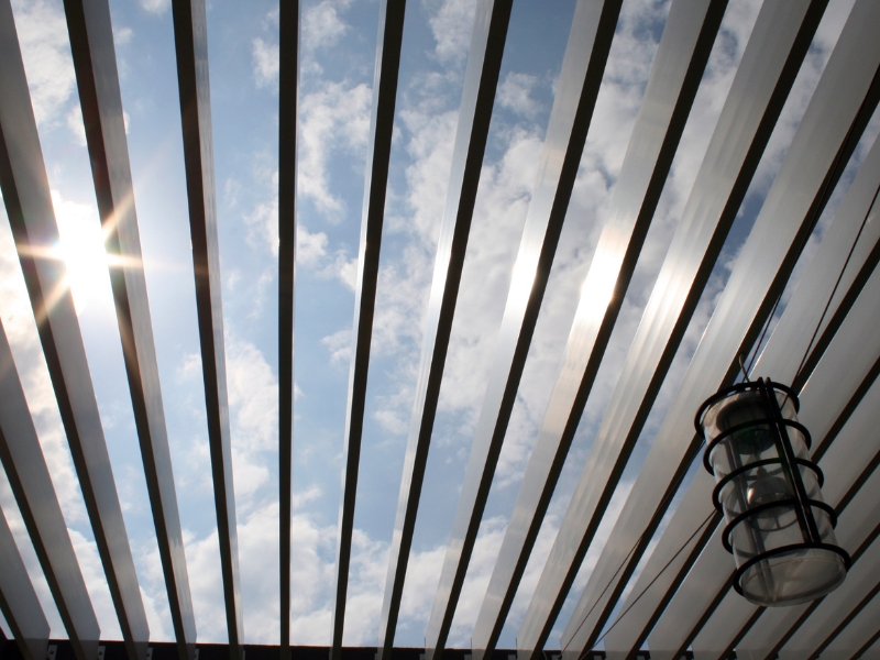 Louvered pergola design plans highlighting roof slats filtering bright sky sunlight.