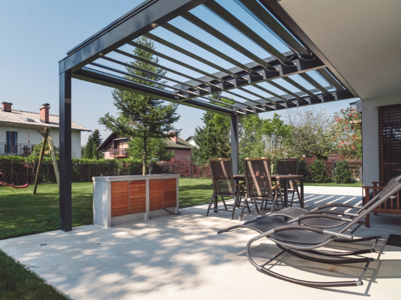 Open steel pergola frame with spaced rafters over patio and lounge chairs in a suburban backyard