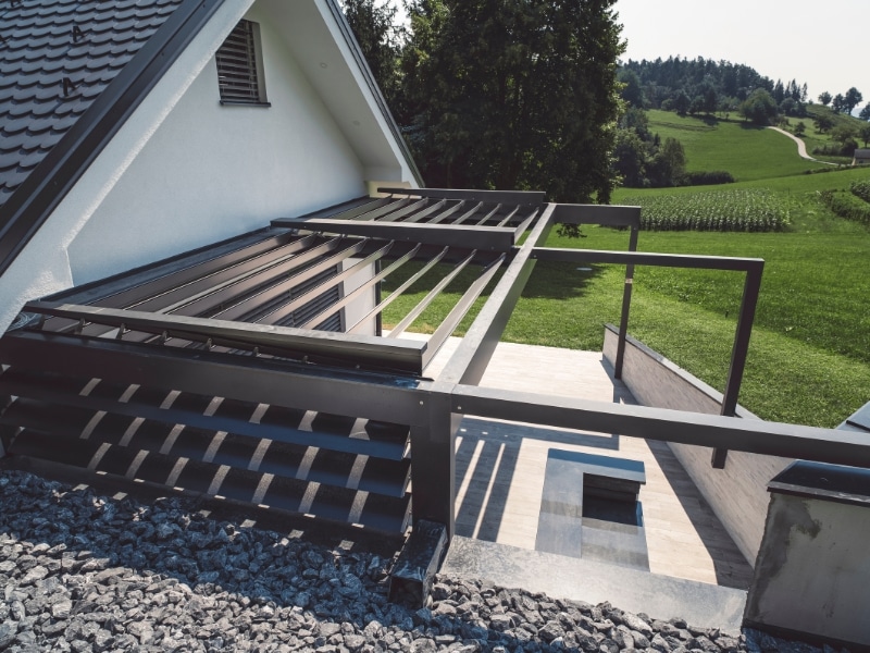 garage pergola designs: modern louvered aluminium pergola frame over side patio beside garage, rural backdrop, under construction.