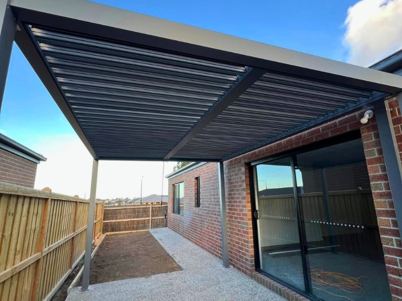 Attaching pergola to house with black louvre roof beside brick wall and concrete patio.