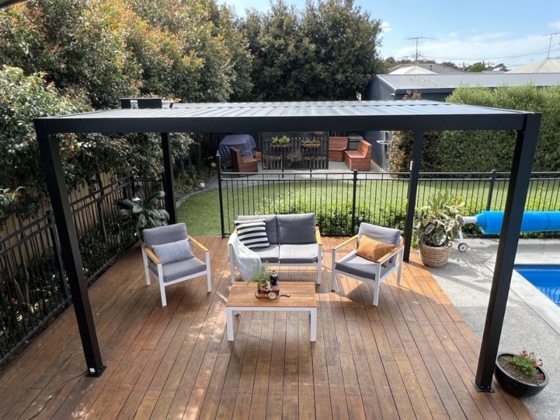 Pergola in front of house on timber deck with outdoor chairs and table.