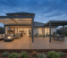 A twilight-view of a modern residential backyard. A wooden deck spans the foreground, featuring a left lounge section with a section sofa, fire pit table, and a contemporary dark-grey aluminum pergola with integrated LED lighting. A central dining area with a table, chairs, and a smaller, unlit pergola sits to the right, adjacent to a gas grill. The house’s illuminated kitchen and living area is visible through large sliding glass doors. The sky is deep blue with soft clouds, and the garden is landscaped with native Australian plants.