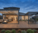 Twilight photo of a modern suburban home's outdoor timber deck, featuring two dark-framed glass-enclosed pergolas. The larger pergola on the left has warm integrated ceiling lighting, louvered roofs, and floor-to-ceiling glass sliding doors enclosing a fully furnished outdoor lounge with a sectional sofa and fire pit. The smaller glass-enclosed pergola on the right houses an outdoor dining area set against a twilight sky and landscaped Australian garden.